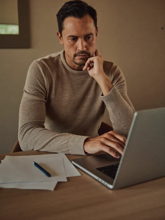A man carefully reviewing paperwork and his laptop at a home desk